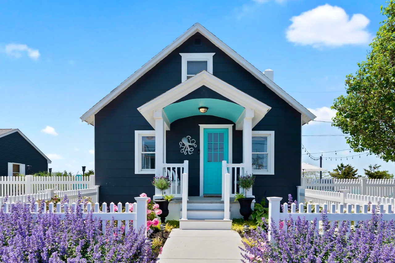 Finished Octopus Cottage with navy siding, a turquoise front door, white fencing and flowering gardens.